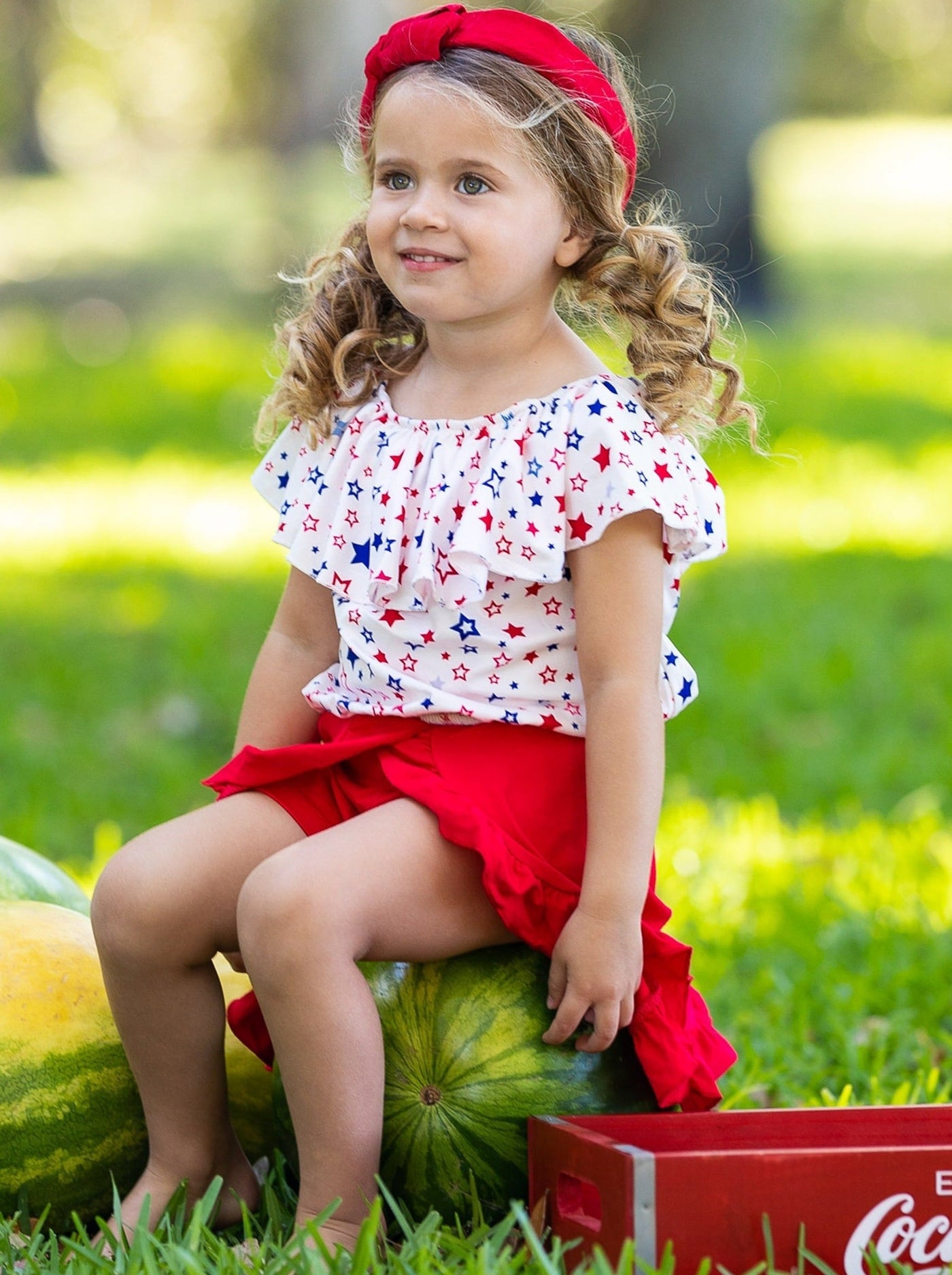 Girls three piece set features a white ruffle top with red and blue star print and red shorts with matching overlay wrap ruffle skirt
