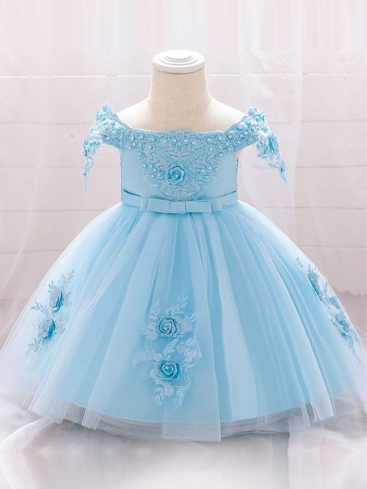 Baby dress has a satin embroidered bodice with pearl details and capped sleeves, a bow belt at the waist, and a tulle skirt with flower applique