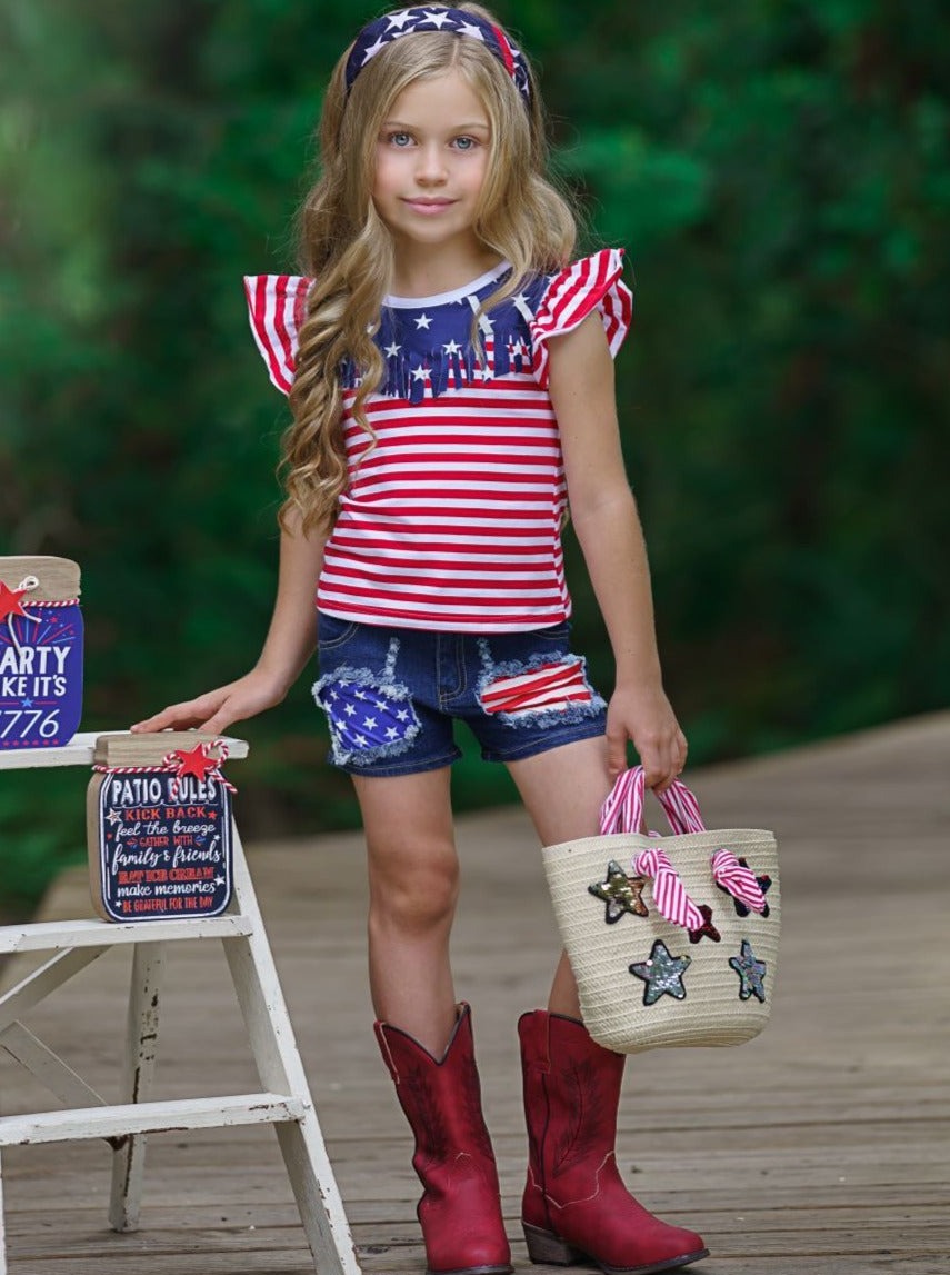 Girls 4th of July set features an American flag print flutter sleeve top with fringe neckline and patched denim shorts with a sash