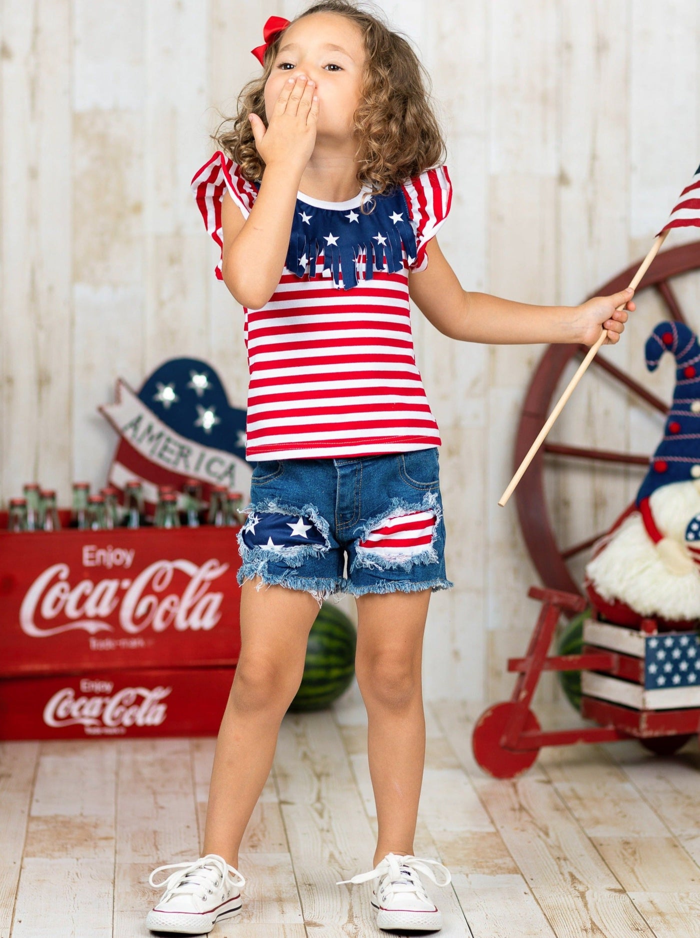 Girls 4th of July set features an American flag print flutter sleeve top with fringe neckline and patched denim shorts with a sash