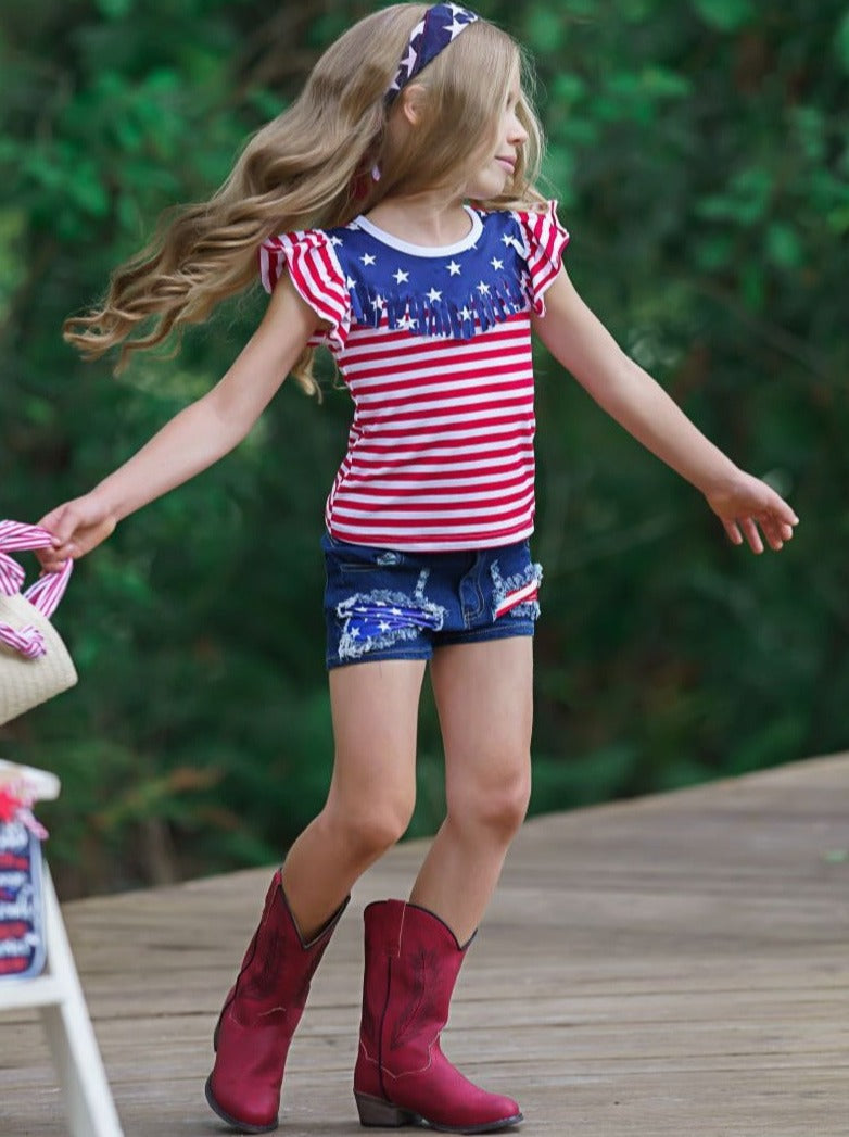 Girls 4th of July set features an American flag print flutter sleeve top with fringe neckline and patched denim shorts with a sash